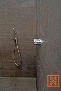 Een strakke, chromen douchekop en kraan zijn gemonteerd op een donkergrijze tegelwand in een moderne badkamer. De doucheslang hangt netjes aan de zijkant, wat zorgt voor een minimalistische en strakke uitstraling.