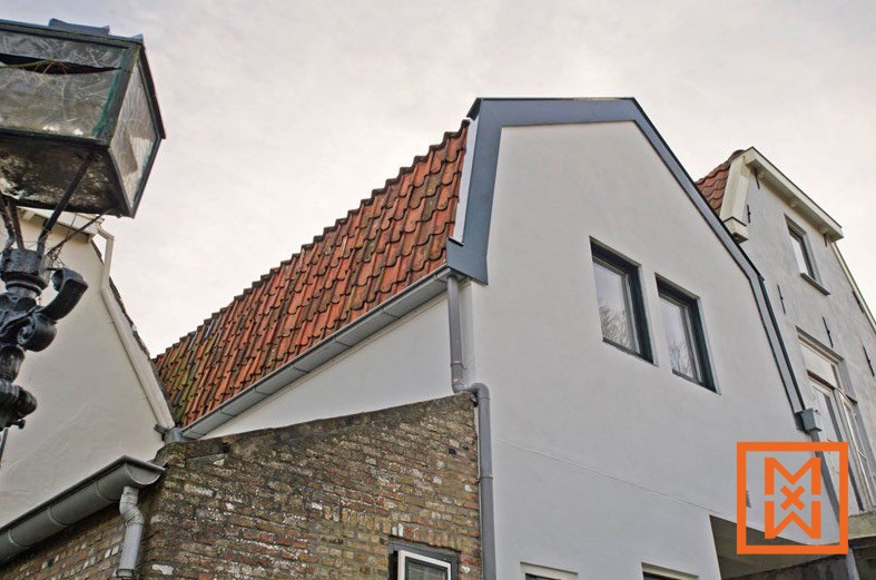 Close-up van de buitenkant van een gerenoveerd huis, met een nieuw gepleisterde grijze muur, een rood pannendak en donkergrijze kozijnen. Een deel van het oudere metselwerk is zichtbaar, wat contrasteert met de moderne gevel. Een oude, beschadigde lantaarnpaal is gedeeltelijk zichtbaar op de voorgrond.
