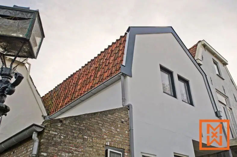 Close-up van de buitenkant van een gerenoveerd huis, met een nieuw gepleisterde grijze muur, een rood pannendak en donkergrijze kozijnen. Een deel van het oudere metselwerk is zichtbaar, wat contrasteert met de moderne gevel. Een oude, beschadigde lantaarnpaal is gedeeltelijk zichtbaar op de voorgrond.