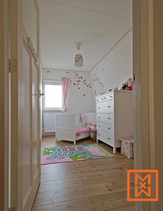 Een lichte, luchtige kinderkamer is voorzien van witte meubels, waaronder een ledikantje en commode, en een speels roze en groen vloerkleed. Vlinders sieren de muren en creëren een speelse sfeer. Natuurlijk licht stroomt binnen via een raam en verlicht de lichte houten vloer.