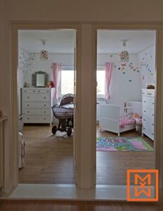 Twee charmante, lichte kinderkamers worden onthuld door open deuren, met twee ledikanten, witte commode en pastelroze accenten. Een rieten wiegje staat op de voorgrond en draagt bij aan de zoete, serene sfeer van deze prachtig ingerichte babykamers.