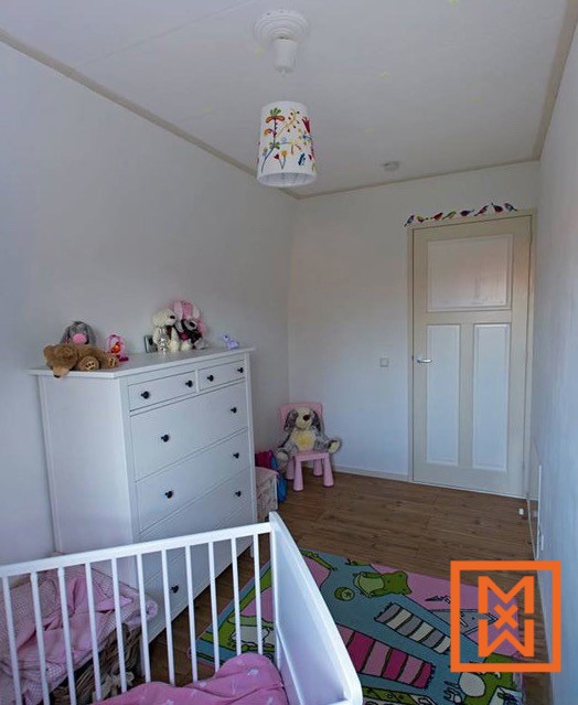Een lichte, vrolijke kinderkamer met een wit ledikantje, een witte commode en een speels vloerkleed met cartoondieren. Knuffels staan op de commode en een stoeltje, wat een gezellige en uitnodigende ruimte creëert voor een baby of peuter. De kamer is geschilderd in een rustgevend wit, met een lichtgekleurde deur en een lampenkap met bloemenmotief die subtiele kleuraccenten toevoegen.