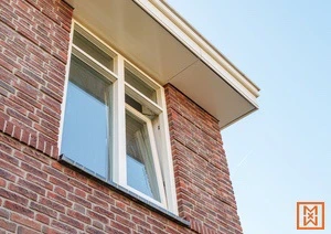 Openstaand draaikiepraam in bakstenen muur onder witte dakrand tegen blauwe lucht.