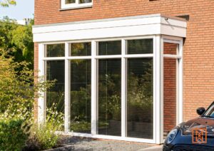 Moderne serre met witte kozijnen aan een bakstenen huis. De grote ramen reflecteren de tuin. Grindpad leidt naar de serre. Een zwarte auto is gedeeltelijk zichtbaar aan de rechterkant.