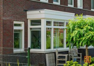 Moderne aanbouw met witte kozijnen aan een bakstenen huis, omringd door groen. Twee houten stoelen staan voor de ramen, wat een uitnodigende sfeer creëert.
