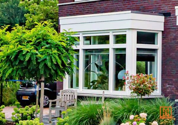 Gezellige erker met witte kozijnen in een bakstenen huis, omgeven door groen en een houten bankje. Een vredig tafereel in de tuin.