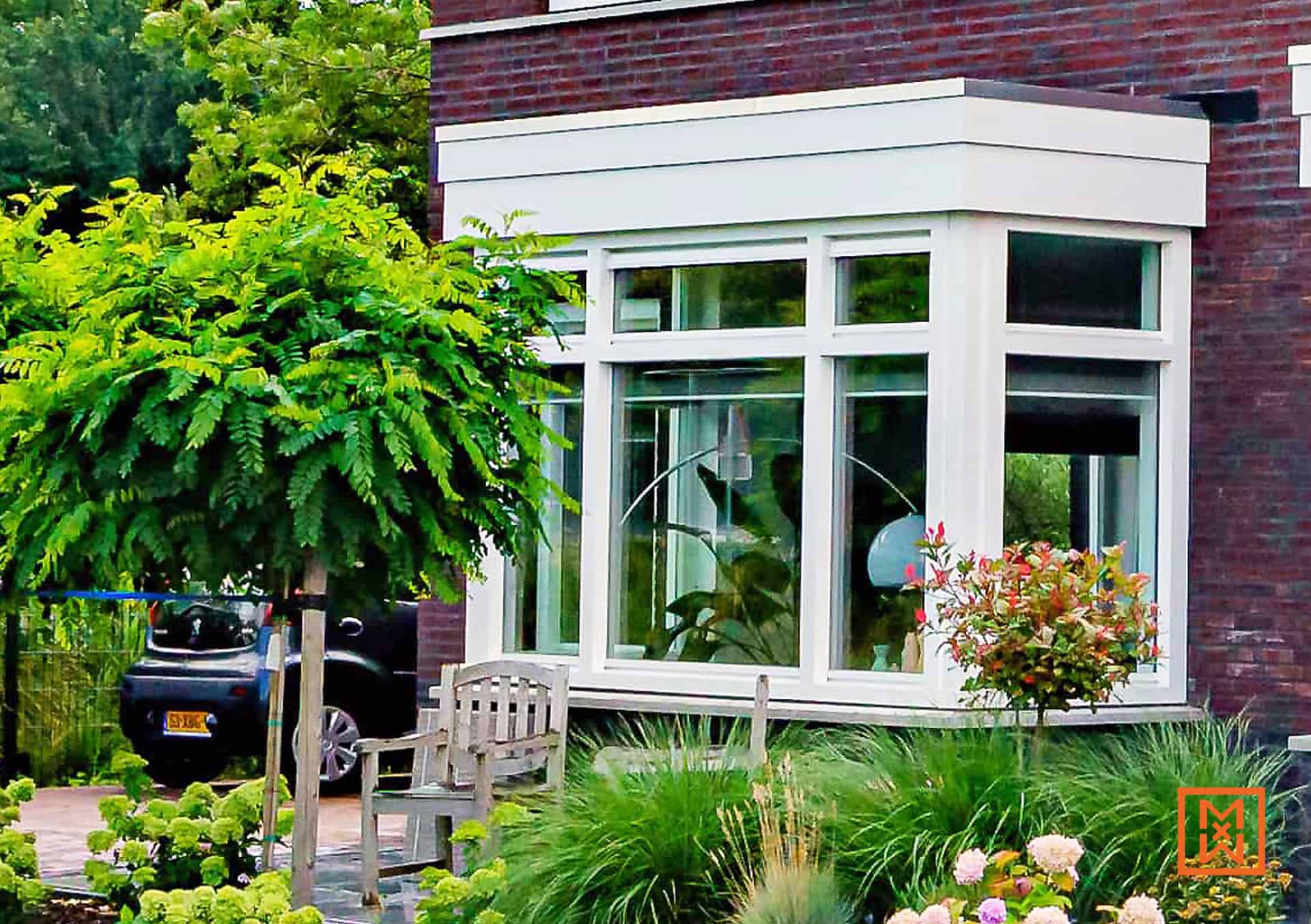 Gezellige erker met witte kozijnen in een bakstenen huis, omgeven door groen en een houten bankje. Een vredig tafereel in de tuin.