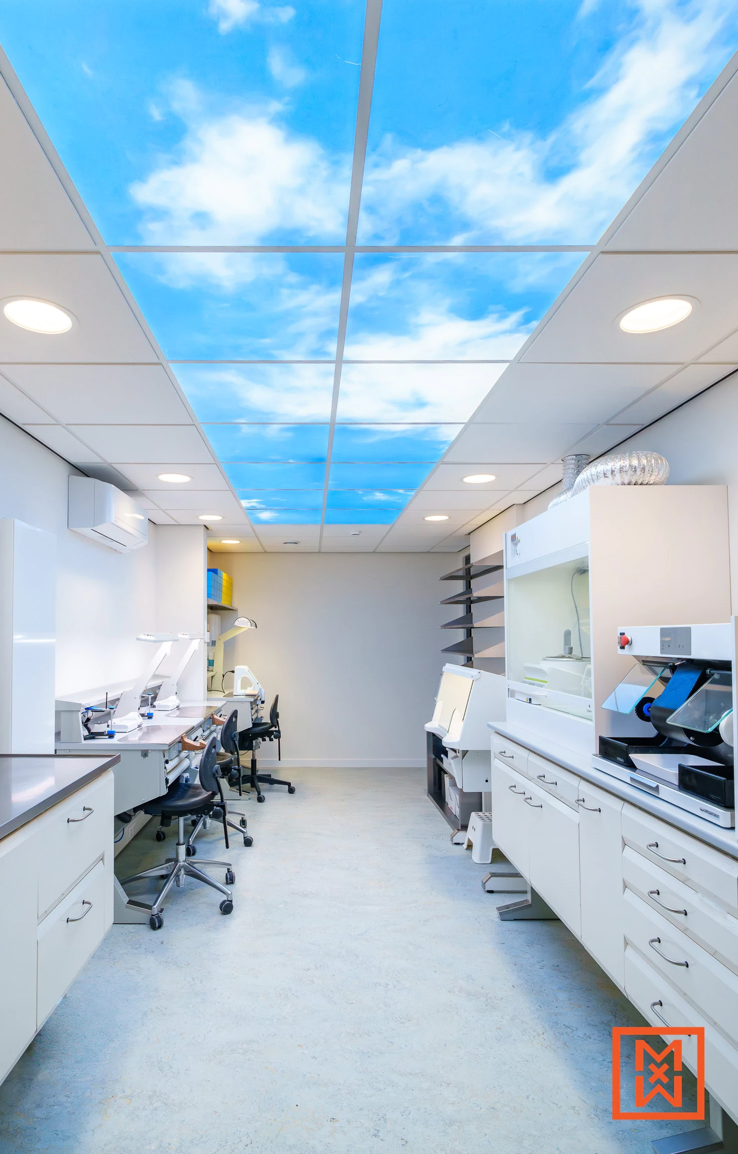 Licht tandtechnisch laboratorium met witte kasten en werkbladen. Een plafond met een blauwe lucht en wolken creëert een ontspannen sfeer.