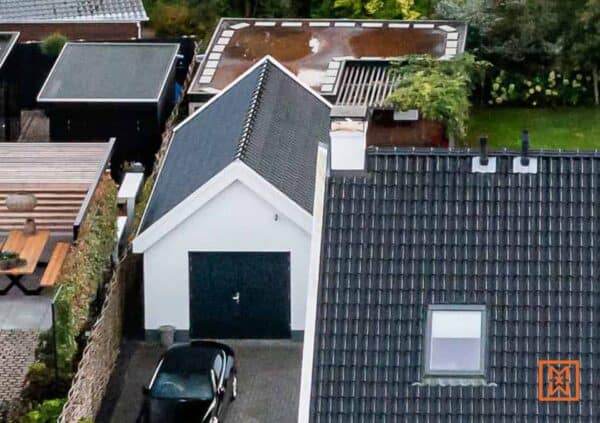 Bovenaanzicht van een garage met een zwarte auto ervoor, omringd door huizen met verschillende dakstijlen, waaronder een plat dak en een puntdak. Grijze bestrating leidt naar de garage.
