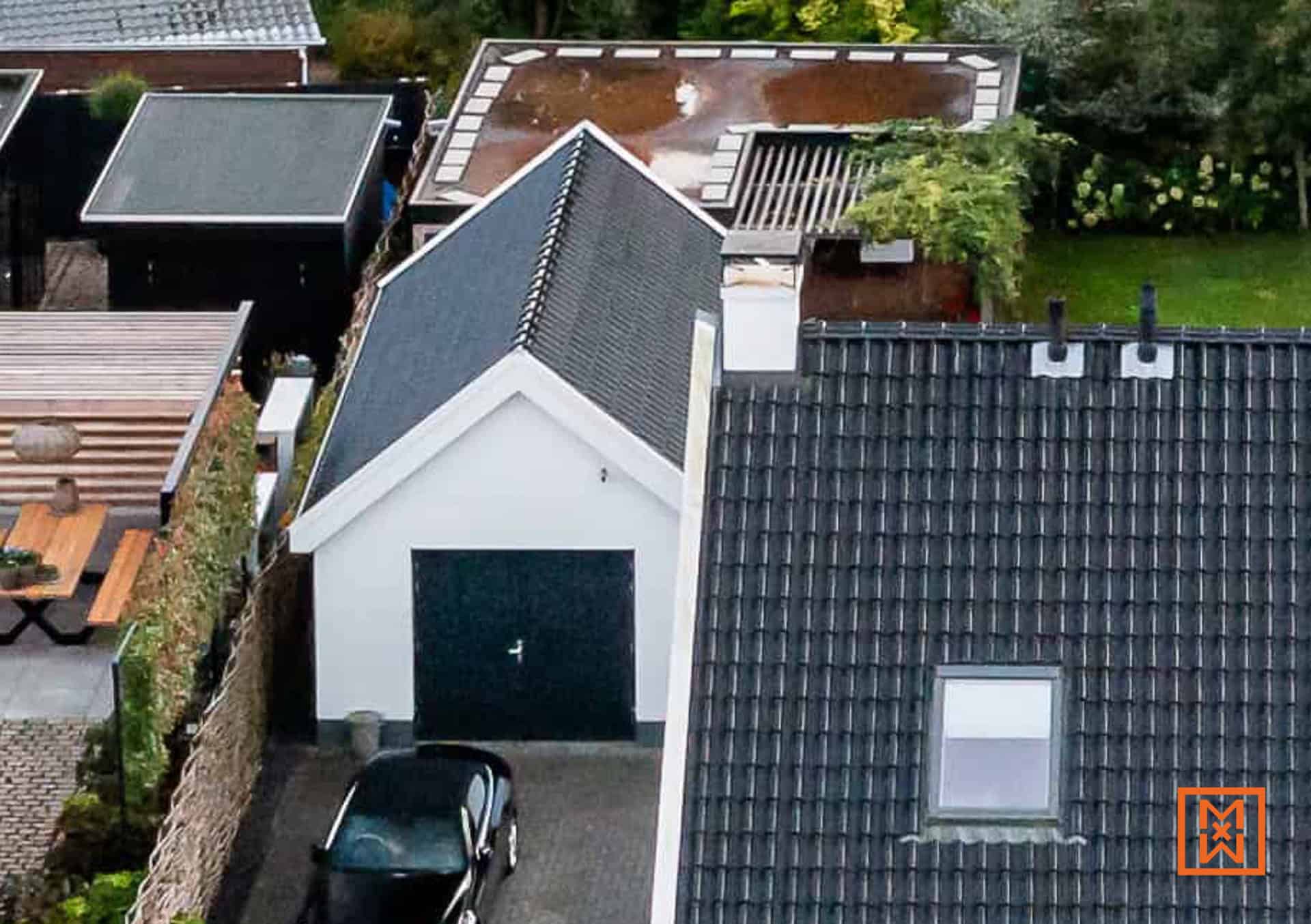 Bovenaanzicht van een garage met een zwarte auto ervoor, omringd door huizen met verschillende dakstijlen, waaronder een plat dak en een puntdak. Grijze bestrating leidt naar de garage.