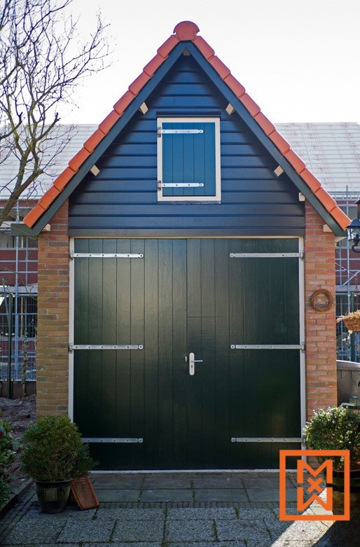 Charmante donkergroene garage in Hollandse stijl met een rood pannendak, met een kleiner donkergroen raam boven de dubbele deuren. Bakstenen zijkanten en een geplaveid stenen pad aan de voorkant maken het plaatje compleet.