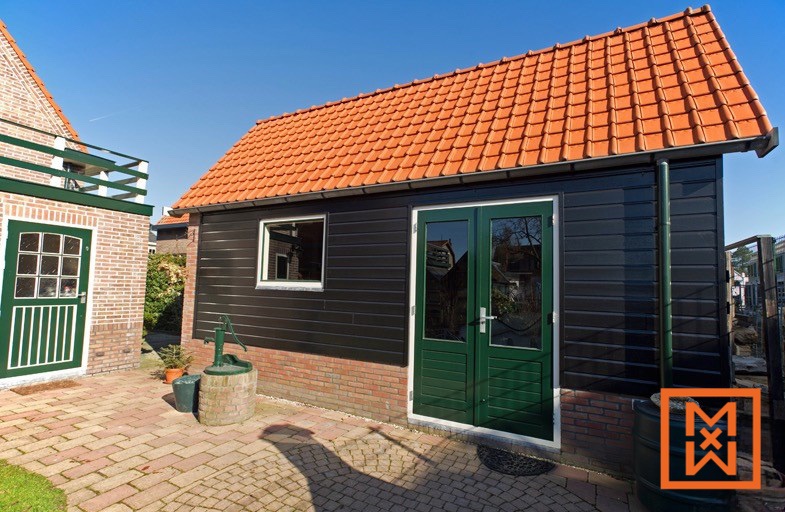 Charmante, zwart beklede tuinschuur met een terracotta dak, dubbele groene deuren en een klein raam. De schuur staat op een met bakstenen geplaveide binnenplaats naast een bakstenen huis, met een waterpomp die een rustieke toets toevoegt.