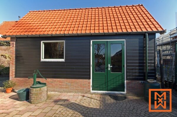 Charmante tuinschuur met zwarte zijkanten, een donkergroene dubbele deur, een oranje pannendak en een klein raam. Ernaast staat een vintage waterpomp op een stenen terras. De zonnige dag zorgt voor een vredige sfeer in de achtertuin.