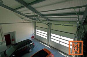 Van bovenaf gezien is het interieur van een ruime garage met twee geparkeerde auto's en drie grote, moderne sectionaaldeuren. De strakke, lichte ruimte heeft een lichtgrijze vloer en witte muren. Het garagedeursysteem is duidelijk zichtbaar.