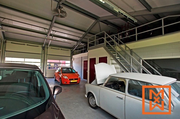 Ruim, modern garage-interieur met hoge plafonds, met drie auto's: een donkerbruine auto op de voorgrond, een feloranje Fiat in het midden en een klassieke lichtgrijze auto met de motorkap omhoog, vlakbij een metalen trap die naar een opslagruimte op de tussenverdieping leidt.
