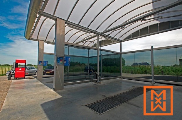 Moderne, overdekte self-service wasstraat met glanzende roestvrijstalen wanden en een polycarbonaat dak. Twee auto's zijn zichtbaar, één geparkeerd en één in de wasstraat, onder een stralend zonnige hemel. Ook een rode betaalkiosk is zichtbaar.