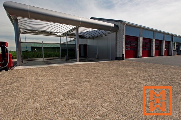 Moderne autowasstraat met een strakke, grijze luifel en rode garagedeuren. Het gebouw is opgetrokken uit lichtgekleurde baksteen en het verharde oppervlak heeft een lichtbruin baksteenpatroon met textuur. Links is een stofzuiger zichtbaar.