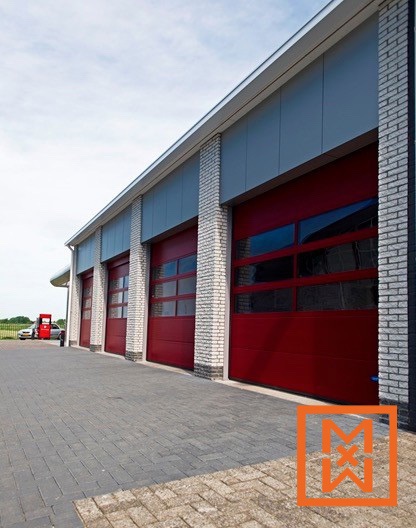 Vier rode sectionaaldeuren met glazen panelen bevinden zich in een modern gebouw met lichtgrijze gevelbekleding en witte bakstenen pilaren. Het gebouw staat op een verhard terrein, met in de verte een klein tankstation zichtbaar onder een gedeeltelijk bewolkte hemel.