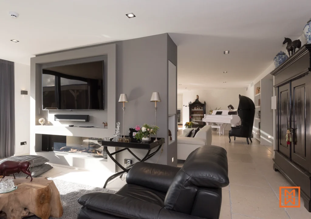 Luxe woonkamer met grijze accenten, een zwarte leren fauteuil en een open haard onder een grote tv. Verderop een witte piano en een doorkijk naar een lichte ruimte. Modern interieur.