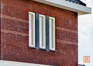 Gevel van een bakstenen huis met een modern raam met drie verticale ramen en donkergrijze stijlen. De bakstenen zijn rood met horizontale witte voegen.