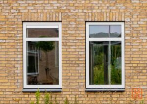 Twee witte ramen in een geel bakstenen gebouw. De ramen reflecteren de omgeving, waardoor een rustieke en huiselijke sfeer ontstaat.
