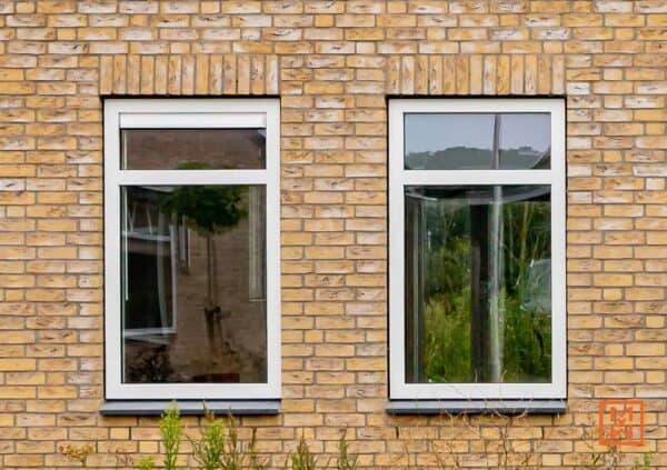 Twee witte ramen in een geel bakstenen gebouw. De ramen reflecteren de omgeving, waardoor een rustieke en huiselijke sfeer ontstaat.