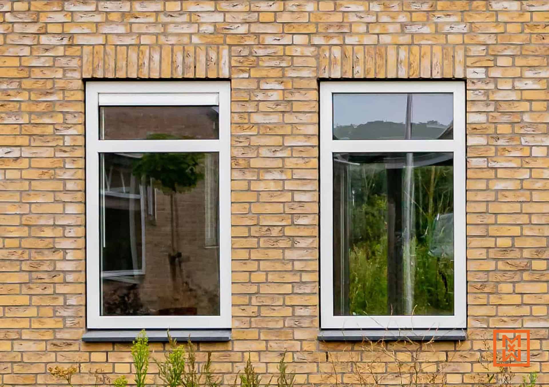 Twee witte ramen in een geel bakstenen gebouw. De ramen reflecteren de omgeving, waardoor een rustieke en huiselijke sfeer ontstaat.