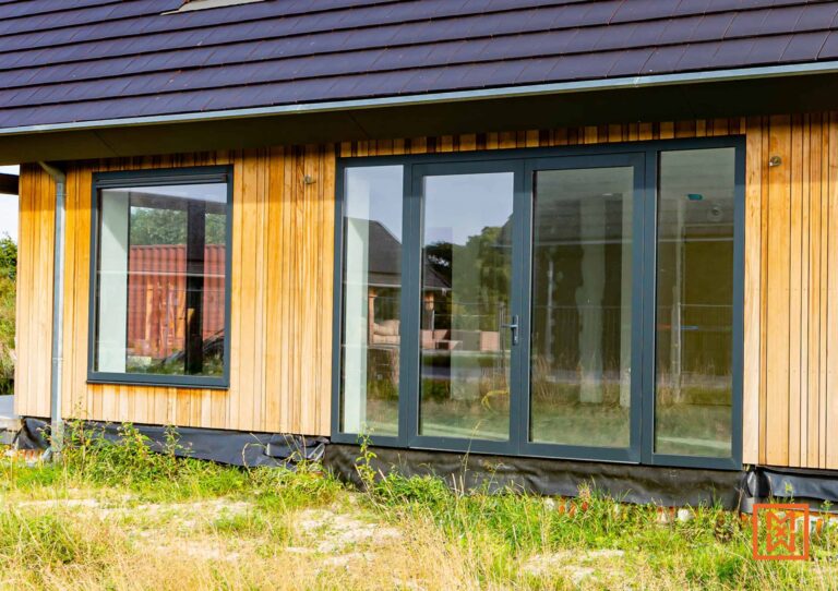 Moderne woning met houten gevelbekleding, grote glazen schuifdeuren en een vierkant raam. De woning heeft een donker dak en bevindt zich in een groene omgeving.