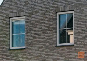 Twee witte ramen in een grijze bakstenen muur. De ramen hebben een kruisverdeling en reflecteren de omgeving. De stenen muur heeft een subtiele textuur.