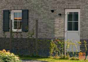 Een grijze bakstenen muur met een wit raam met donkergrijze luiken en een witte deur. Een metalen hek met begroeiing staat voor de muur.