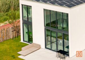Modern huis met zonnepanelen op het dak. Grote ramen met zwarte kozijnen, terras met loungebank en eettafel. Groene tuin met houten schutting. Duurzame architectuur.
