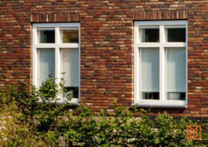 Twee witte ramen in een bakstenen muur, gedeeltelijk bedekt door groene struiken. De ramen hebben een klassieke indeling met roeden. De warme tinten van de bakstenen contrasteren met het wit van de ramen.