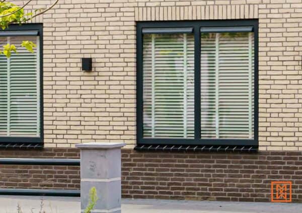 Gevel met ramen met witte jaloezieën en donkergrijze kozijnen. De gevel is van gele baksteen, en er is een plint van bruine baksteen. Een klein vierkant zwart lampje hangt aan de muur.