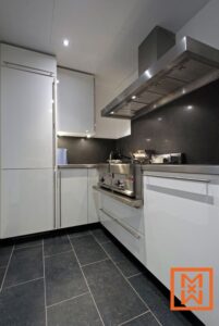 Strakke, moderne keuken met witte kasten, roestvrijstalen apparatuur en donkergrijze werkbladen. Een ingebouwde afzuigkap en een donkere stenen achterwand completeren het eigentijdse ontwerp. De donkergrijze vloertegels dragen bij aan de verfijnde esthetiek.