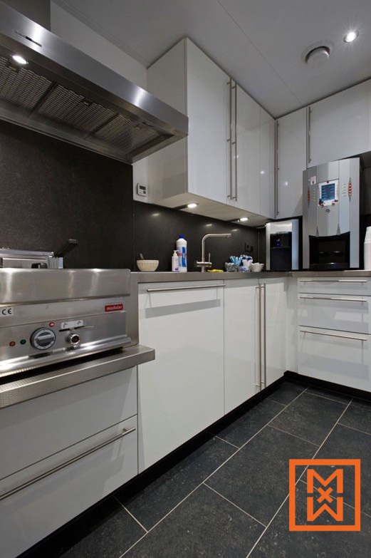 Moderne, witte professionele keuken met roestvrijstalen apparatuur, waaronder een afzuigkap, frituurpan en koffiezetapparaat. Donkergrijze stenen aanrechtbladen en vloeren completeren het strakke ontwerp.