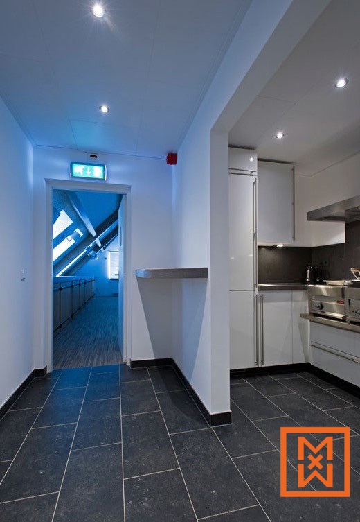 Een moderne, strakke hal met donkergrijze tegelvloer leidt naar een lichte kantoorruimte. Rechts is een minimalistische keuken met witte kasten en roestvrijstalen apparatuur te zien. Inbouwspots verlichten het geheel.