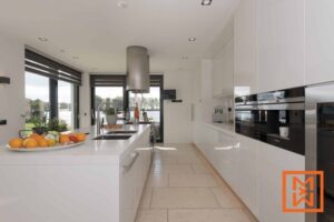 Moderne witte keuken met een groot kookeiland, roestvrijstalen apparatuur en grote ramen met uitzicht op een rustig meer. Het kookeiland is voorzien van een schaal met vers fruit. Natuurlijk licht stroomt de ruimte binnen en creëert een lichte en luchtige sfeer.