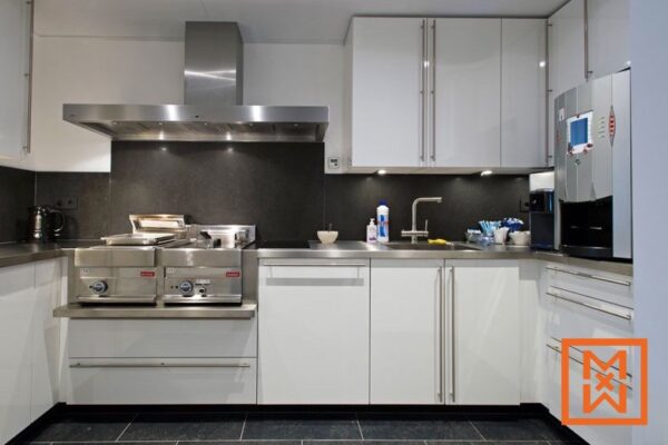 Moderne witte keuken met roestvrijstalen apparatuur, waaronder een afzuigkap, dubbele frituurpannen en een ingebouwd koffiezetapparaat. Een donkergrijze achterwand en strakke werkbladen maken het ontwerp compleet.