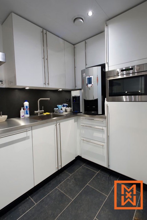 Moderne, witte keuken met roestvrijstalen aanrechtbladen en apparatuur. Inclusief een ingebouwde koffiemachine, magnetron en strakke kasten. De donkergrijze vloertegels zorgen voor een verfijnd contrast.