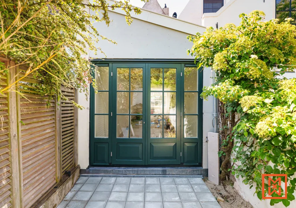Groene tuindeuren met glas in een lichte gevel, omringd door groen. Grijze tegels leiden naar de deuren. Een serene en groene buitenruimte.