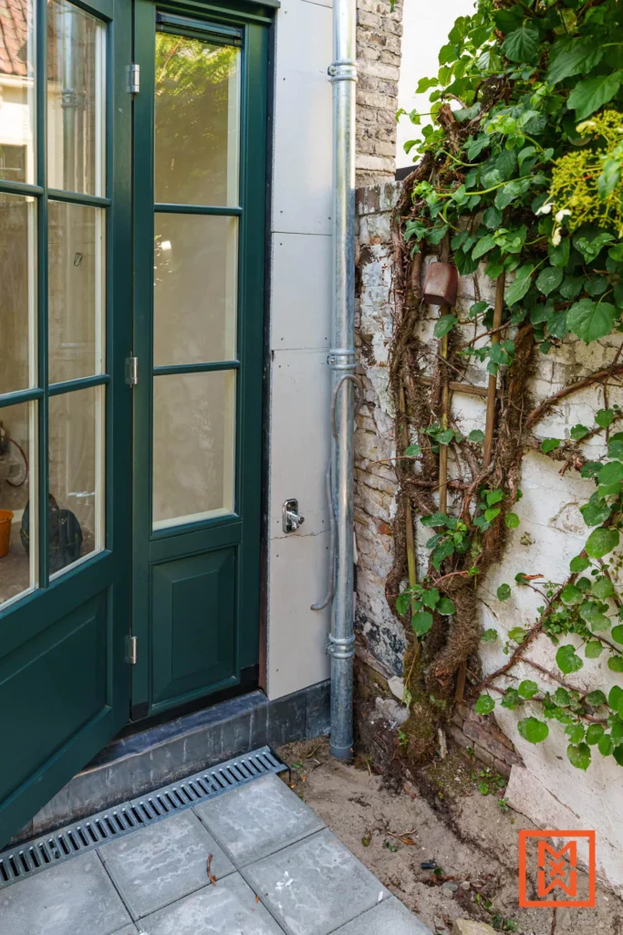 Groene tuindeur naast een witte muur begroeid met klimop. Een zinken regenpijp loopt langs de muur. Bestrating leidt naar de deur. Sfeervolle buitenscène.
