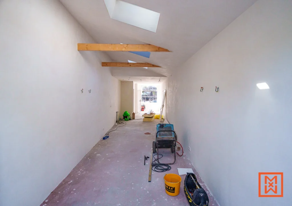 Interieur van een kamer in renovatie met wit gestucte muren, houten balken en een dakraam. Gereedschap en materialen liggen verspreid op de vloer.