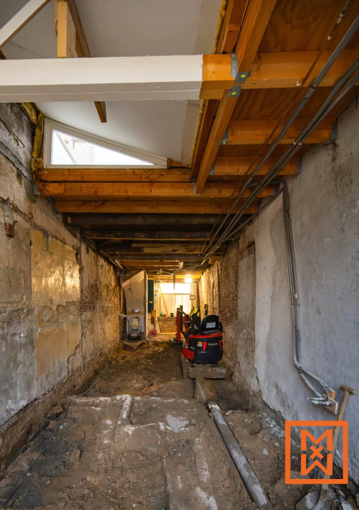 Een kelder in aanbouw met blootgelegde leidingen en een kleine graafmachine. Een driehoekig raam laat licht binnen in de donkere ruimte, waar de muren ruw en onafgewerkt zijn.