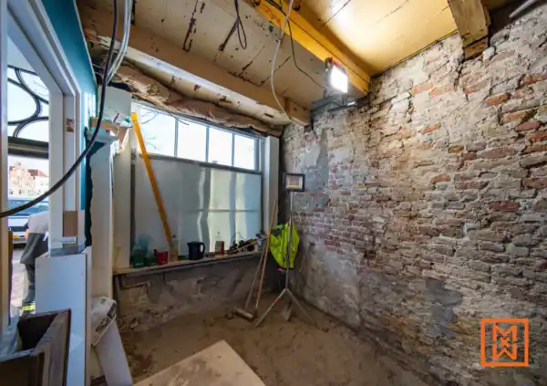 Interieur van een kamer in renovatie met zichtbare bakstenen muur, opengebroken plafond en een raam met veel licht. Er staat bouwgereedschap en een veiligheidshesje.
