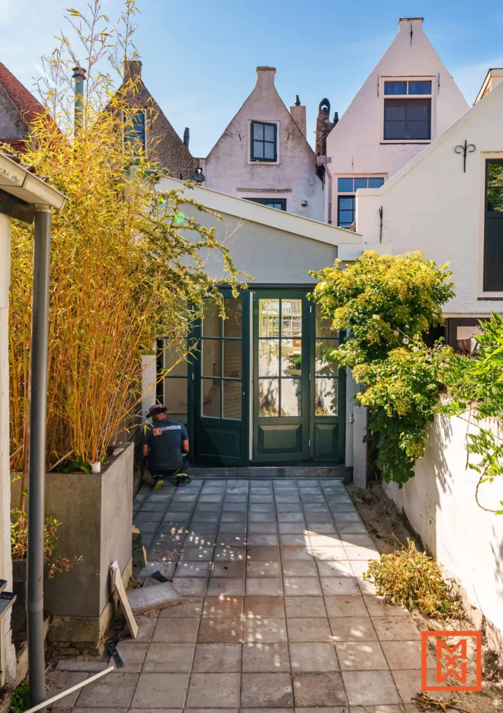 Een groene deur met glas in een betegelde tuin, omringd door bamboe en planten. Op de achtergrond typische Nederlandse gevels onder een blauwe lucht.