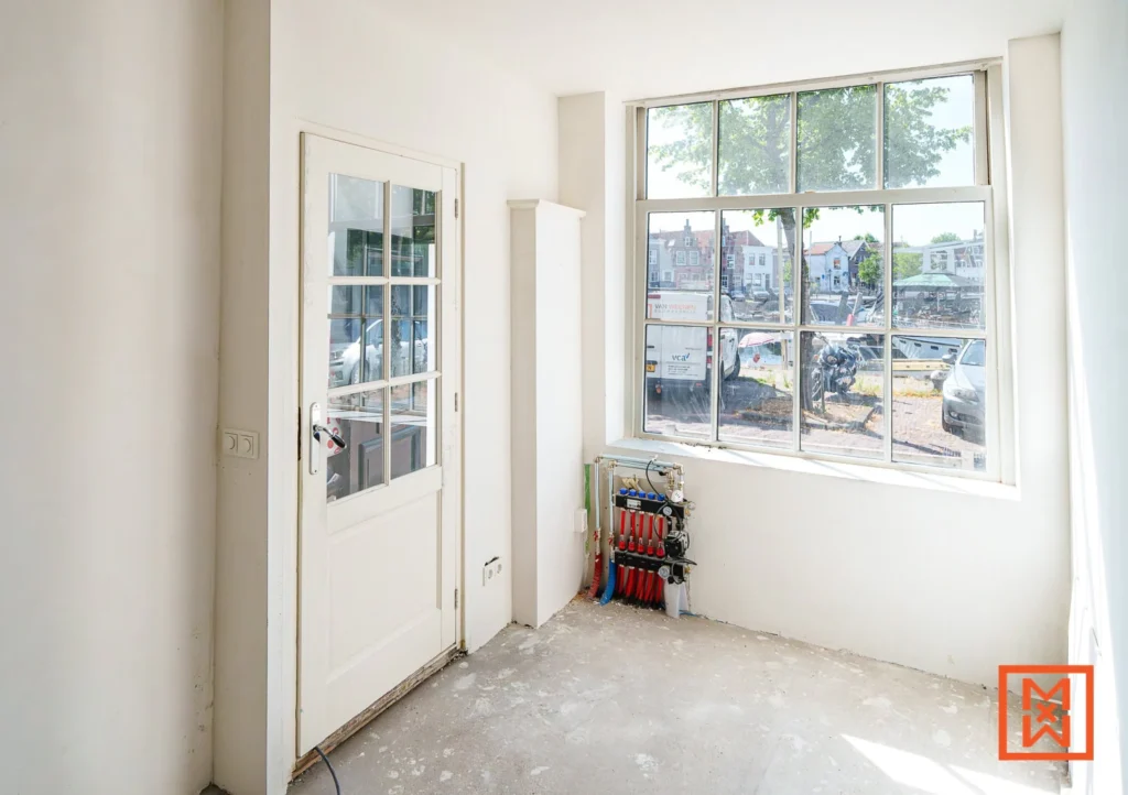 Lichte kamer met een witte deur met glas, een groot raam met uitzicht op een straat in een Nederlandse stad en een verwarmingssysteem.