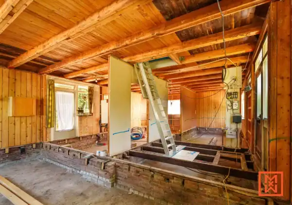 Interieur van een huis in renovatie: houten balken plafond, kale muren, open vloer met bakstenen fundering en een ladder die naar boven leidt. Rommelige maar veelbelovende renovatie.