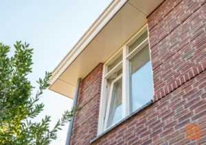Modern bakstenen huis met wit raamkozijn en dakoverstek tegen een blauwe lucht en groen gebladerte.