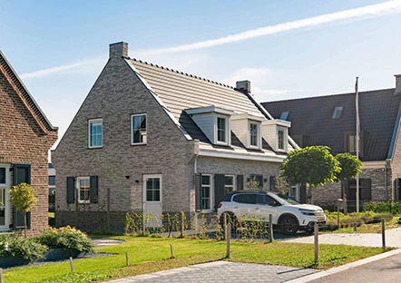 Modern huis met grijze bakstenen, witte kozijnen en een donker dak. Een witte auto staat geparkeerd voor het huis. Groene tuin met jonge boompjes.