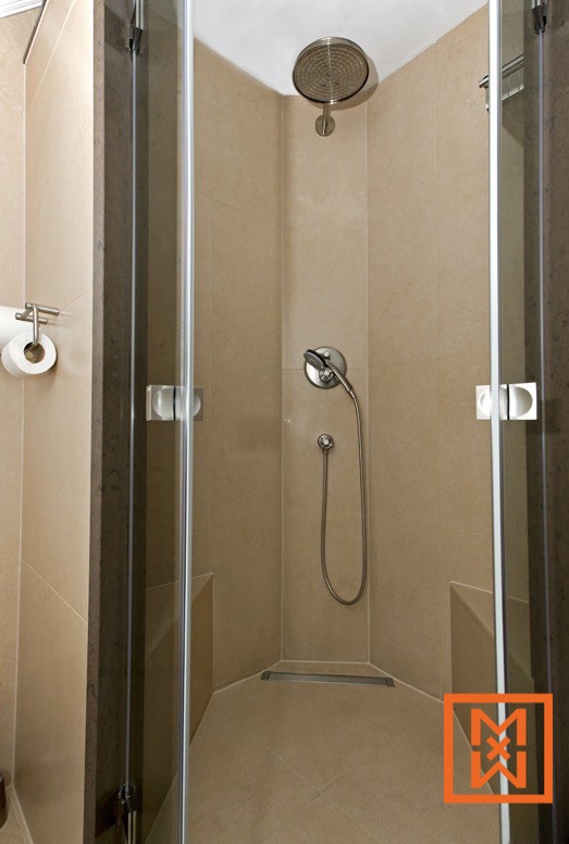 Moderne inloopdouche met beige tegels, glazen deuren, ingebouwde zitting en een regendouchekop. De douche is voorzien van strakke chromen armaturen en een lineaire afvoer.